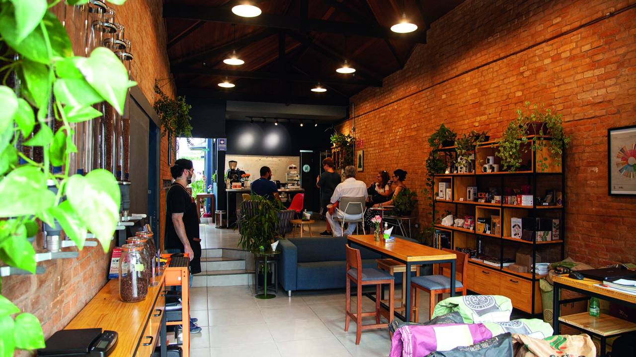 Ambiente do café decorado por altas banquetas de madeira, estofado de cor cinza, prateleiras e mais