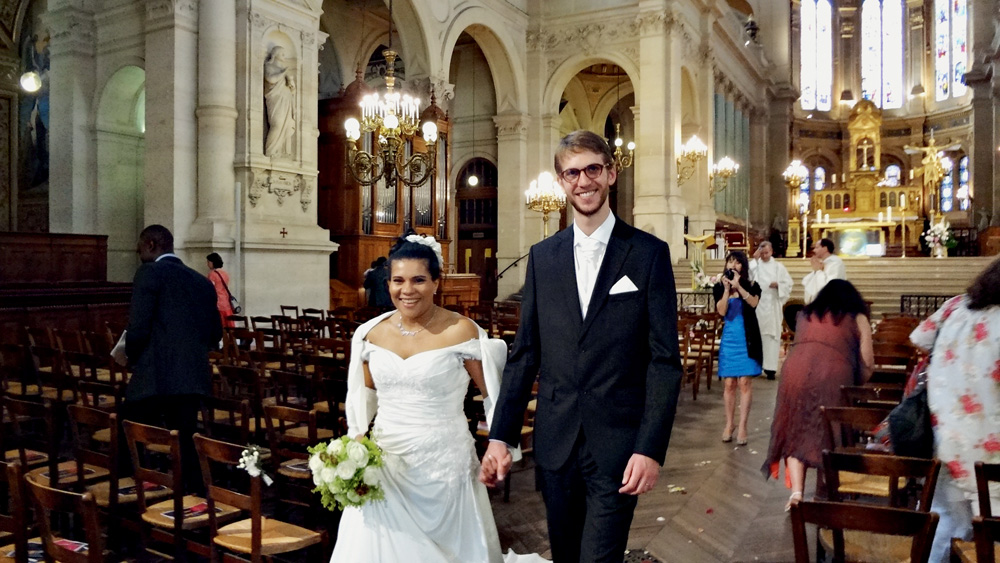 DSC04182 (1).JPG (1) Luciane e Paul no dia de seu casamento. Ele veste um terno preto e óculos. Ela veste um longo vestido bordado com uma coroa. O buquê que ela segura tem rosas brancas.