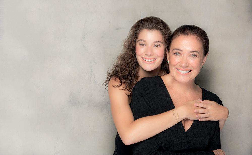 Lilia e Giulia atual Giulia, uma mulher branca, de olhos e cabelos castanhos, abraça a mãe, Lilia, por trás. Ambas sorriem.