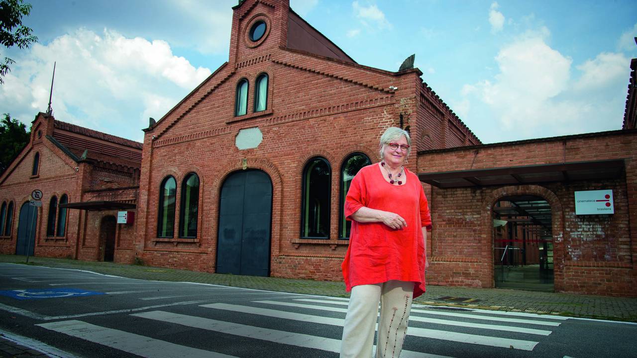 Dora aparece em frente à sede da Cinemateca. Ela é uma mulher idosa, branca e de cabelos grisalhos. Usa óculos, uma calça branca e uma blusa coral até os joelhos.