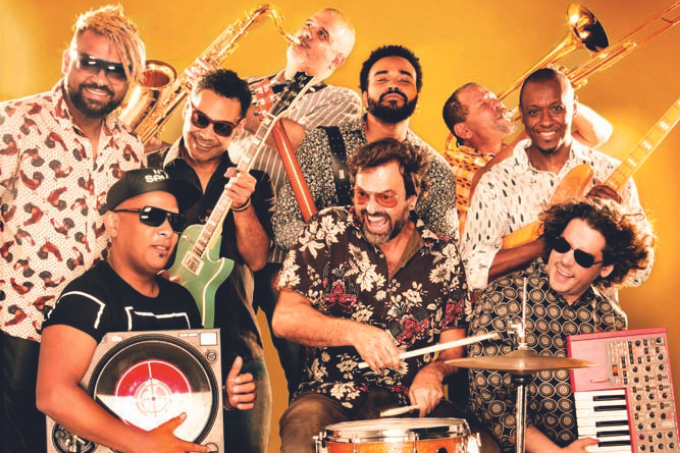 Imagem mostra diversas pessoas com instrumentos em frente a fundo amarelo.