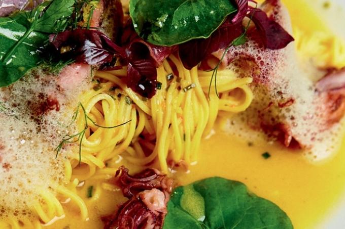 Capellini servido em prato branco com caldo de cor amarela, espuma, lulinhas e folhas verdes e de cor roxa