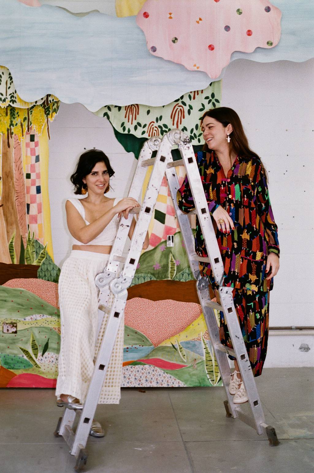 Fernanda Feher e Betina de Luca (Marina Nacamuli-Divulgação) Fernanda Feher e Betina de Luca posam penduradas em escada de alumínio. Fernanda, à esquerda, olha para a câmera, e Betina sorri voltada para Fernanda. Uma veste roupas brancas, a outra veste um conjunto escuro estampado nas cores preto e vermelho.