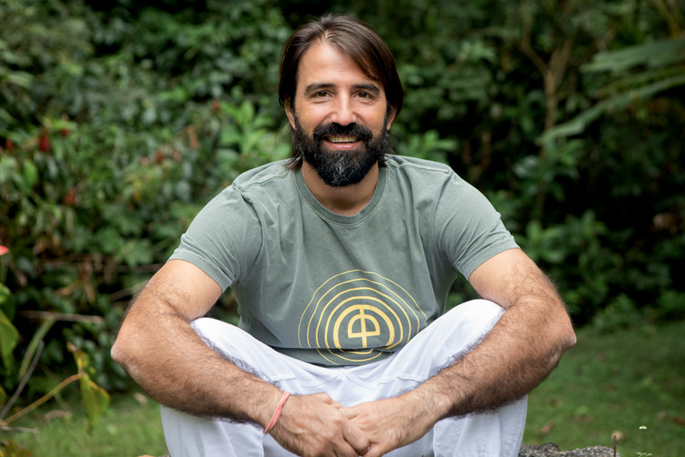 Fernando é um homem branco, magro, de cabelos escuros e compridos e barba. Ele está sentado na grama, em uma área verde, e veste uma camiseta verde e uma calça branca.