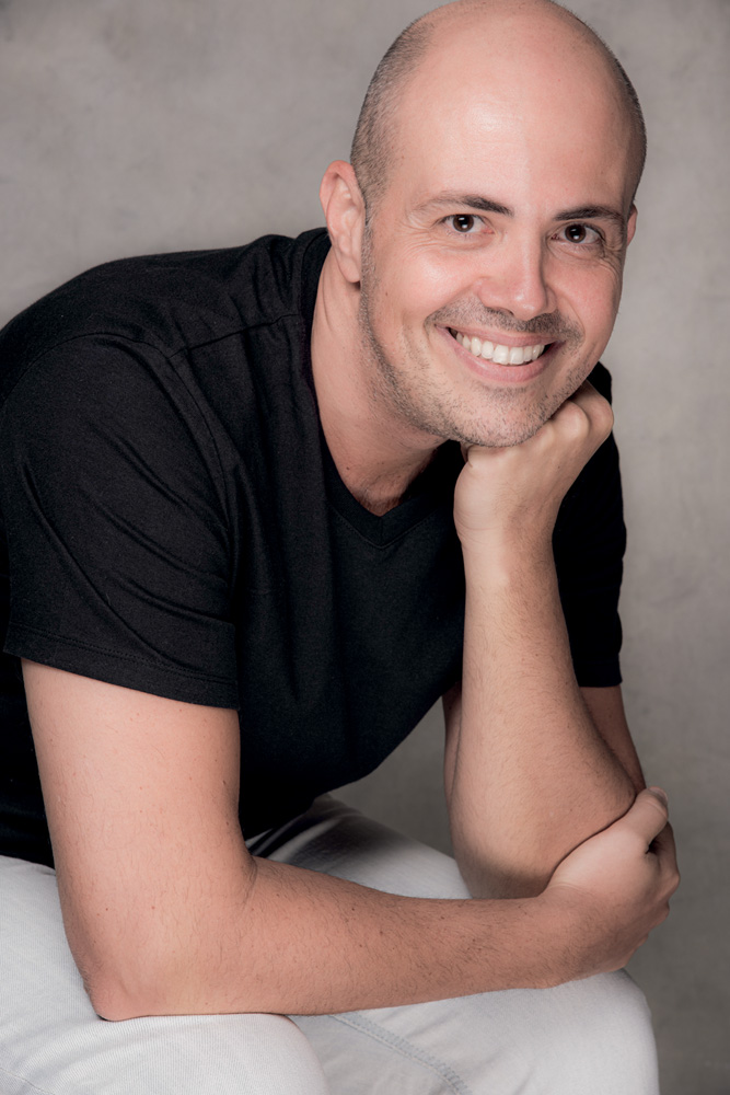 GUSTAVO PINHEIRO COR (6) CREDITO PINO GOMES.jpg Gustavo é um homem branco e careca. Ele usa uma camiseta preta e uma calça branca e sorri para a câmera com os braços apoiados no colo. Ele está sentado