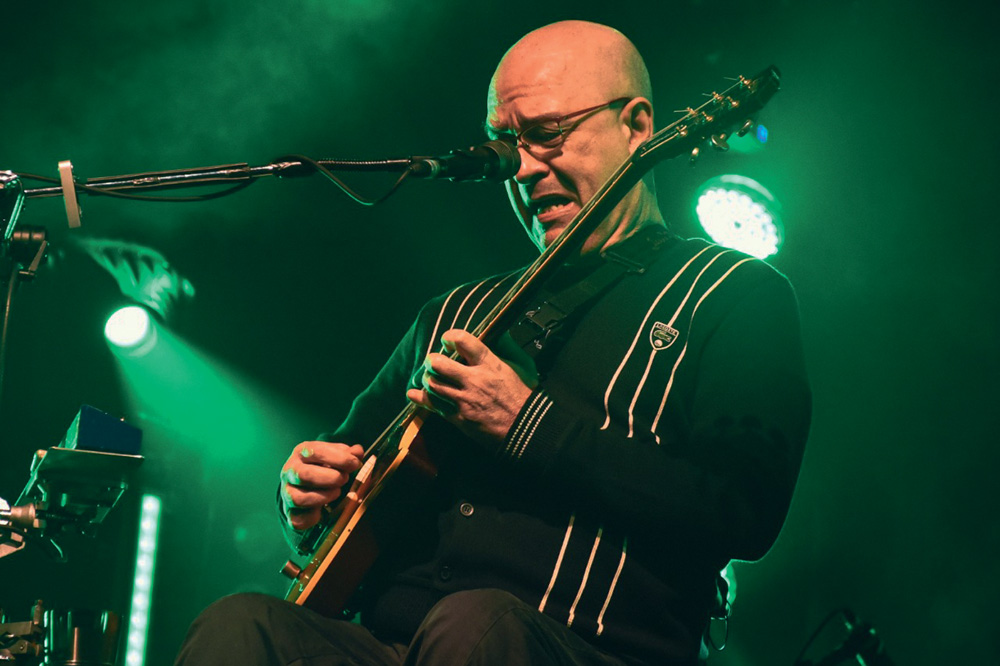 Imagem mostra homem careca sentado tocando guitarra. Ao fundo, um holofote verde.