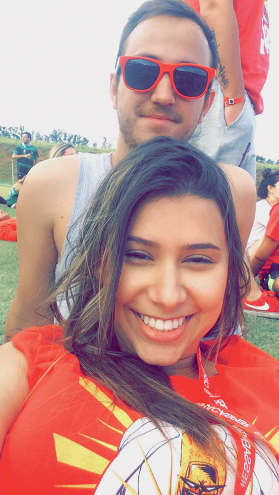 IMG_0729 (1).JPG Beatriz faz uma selfie com Felipe atrás. Ele usa um óculos de sol e ela uma blusa vermelha. Eles estão sentados em um campo gramado