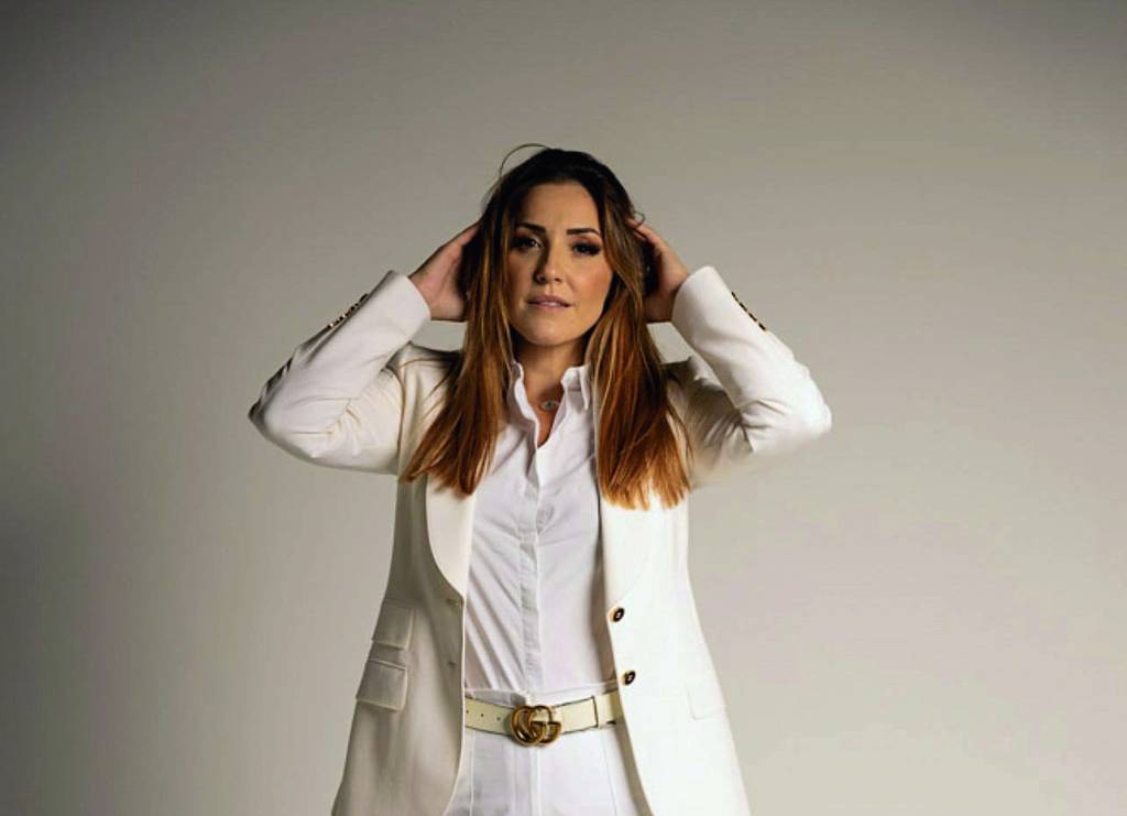 IMG_7974.JPG Thais é uma mulher branca, de cabelos e olhos castanhos. Ela usa uma camiseta, uma calca e um blazer pretos. Da cintura para cima, ela é fotografada em um fundo branco, com as mãos na cabeça