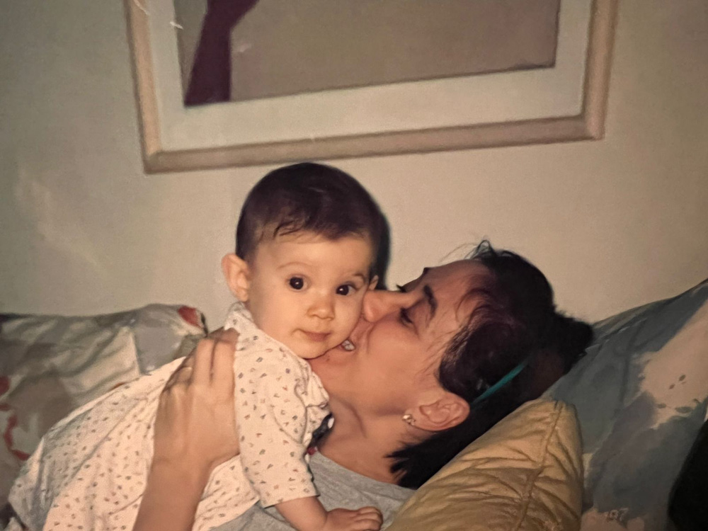 Lilia e Giuliabb Lilia segura Giulia, uma bebê de olhos e cabelos escuros, enquanto dá um beijo em sua bochecha. Elas estão deitas em um sofá e foto é antiga