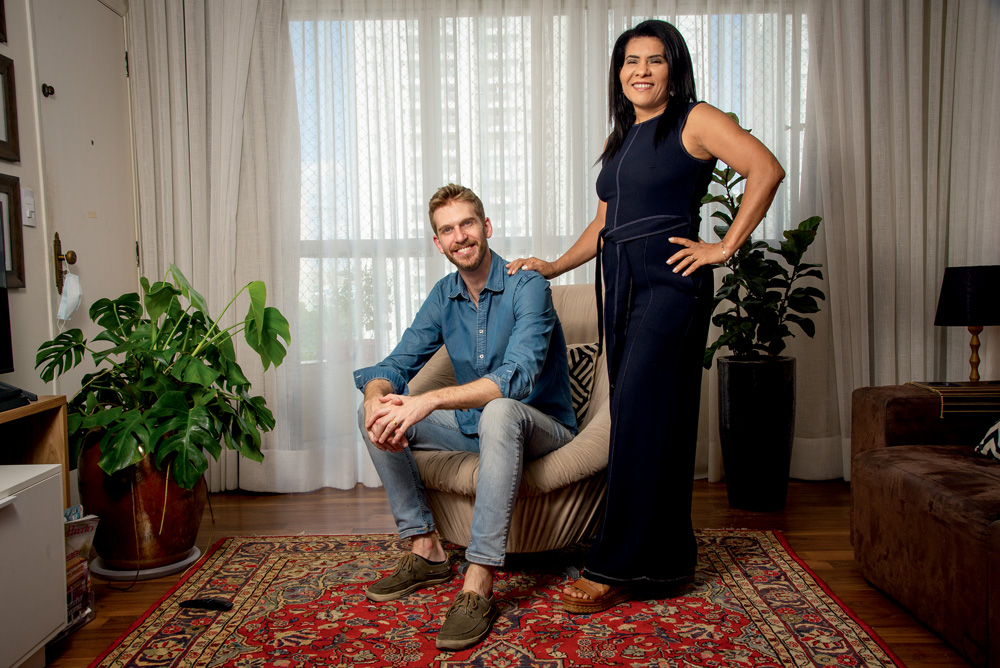 Paul é um homem branco, magro, loiro e de olhos azuis. Ele está sentado em uma poltrona enquanto sua esposa, Luciane, uma mulher negra, de cabelos escuros e crespos, apoia uma das mãos em seu ombro.