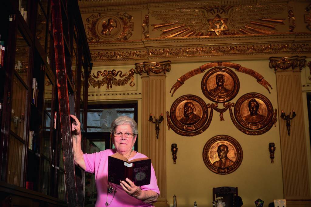 Suely é uma senhora idosa, branca, de cabelos curtos e grisalhos. Ela usa um suéter rosa e posa ao lado de uma estante de madeira, com um livro na mão. A construção é antiga e, ao fundo, é possível ver quadros com fotos de quatro homens banhadas em ouro.