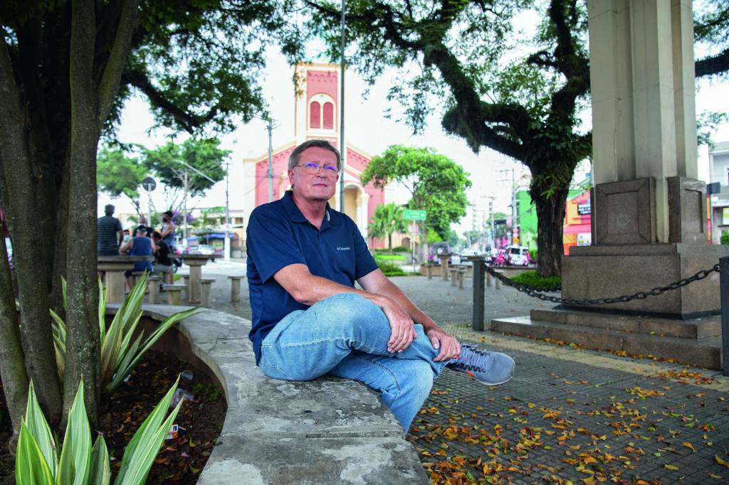 Vila Zelina – Victor Gers Imagem mostra homem de calça jeans e camisa polo azul marinho sentado em praça. Ao fundo, a fachada de uma igreja.