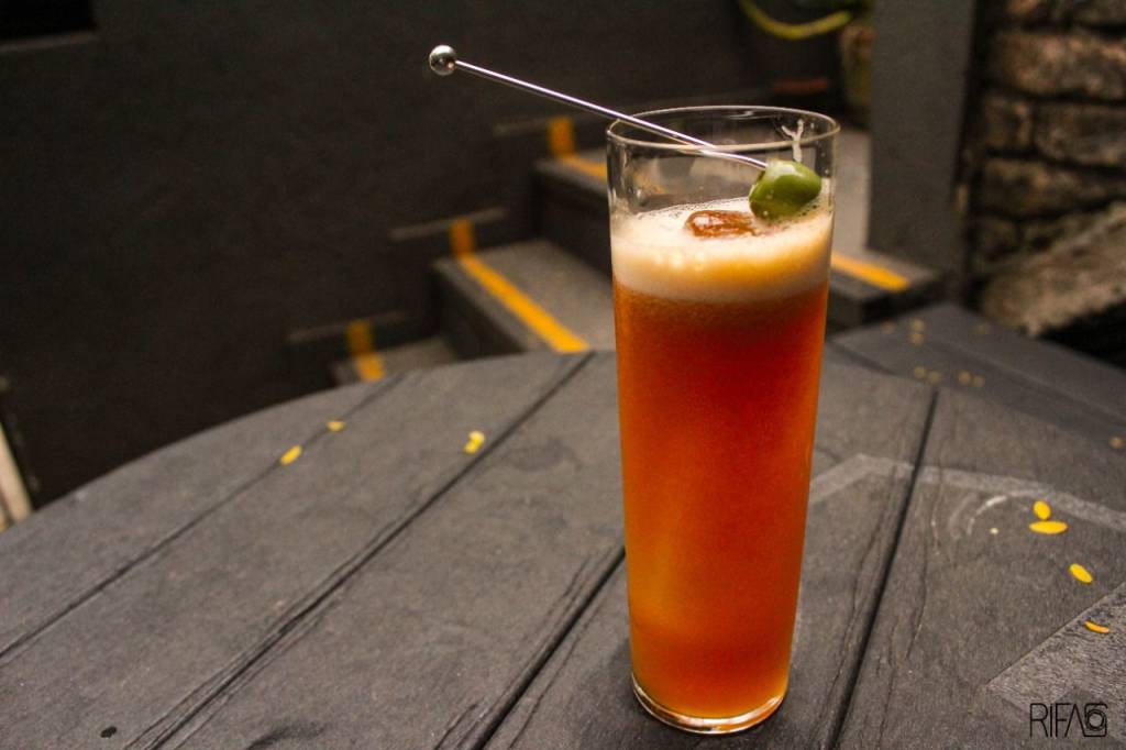 Espaço 13 Drinque de cor alaranjada servido em copo longo com guarnição de azeitona verde