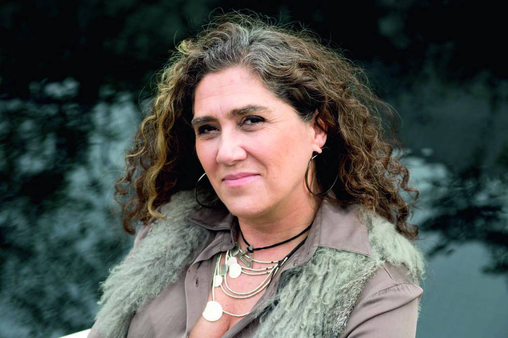 anna muylaert – credito arquivo pessoal.jpg Anna é uma mulher branca, magra, de cabelos castanhos e cacheados. Ela posa para a foto, em que aparece do ombro para cima, e usa colares e um colete cinza de pelos.
