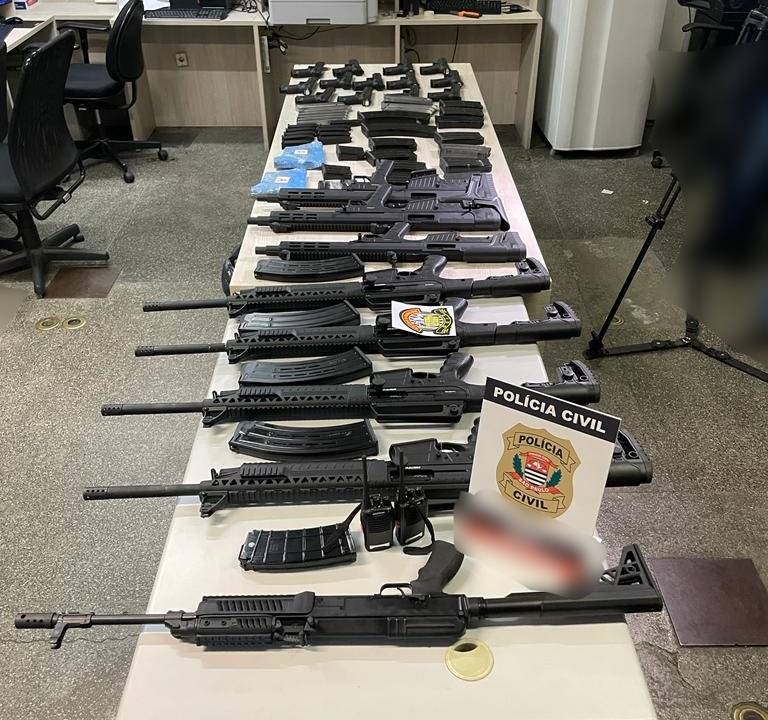 Imagem mostra dezenas de fuzis e pistolas dispostos em mesa de delegacia
