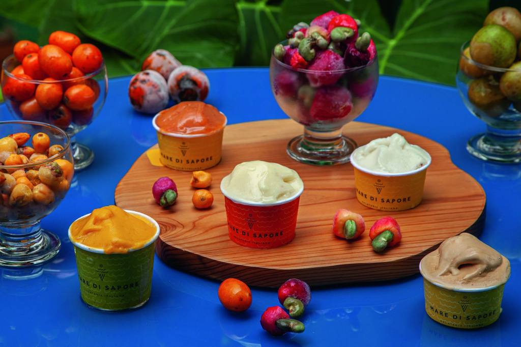 Taça de vidro com sorvete sobre base redonda de madeira, com pedaço de goiabada, pedaço de queijo e pequena faca ao lado, sobre mesa de madeira branca