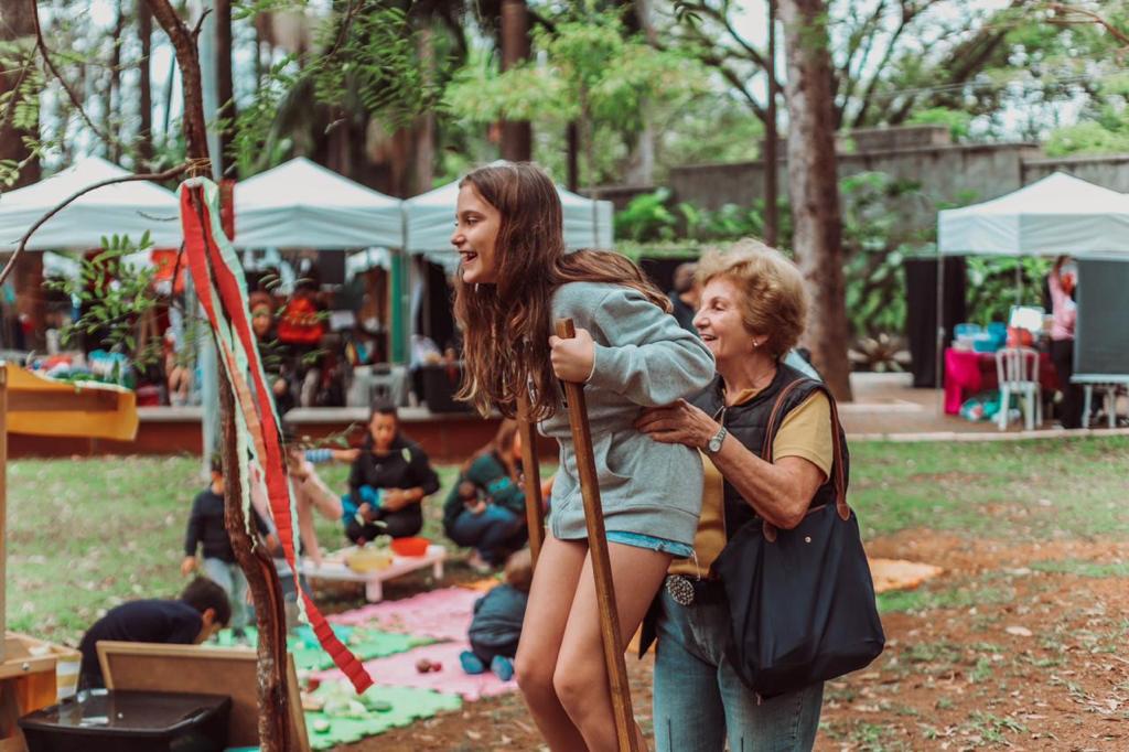 Uma senhora idosa brinca com uma menina em uma feira ao ar livre. Ambas são brancas, de cabelos castanhos. A senhora tem cabelos curtos e usa camiseta e calça jeans. A memina usa um moletom azul claro