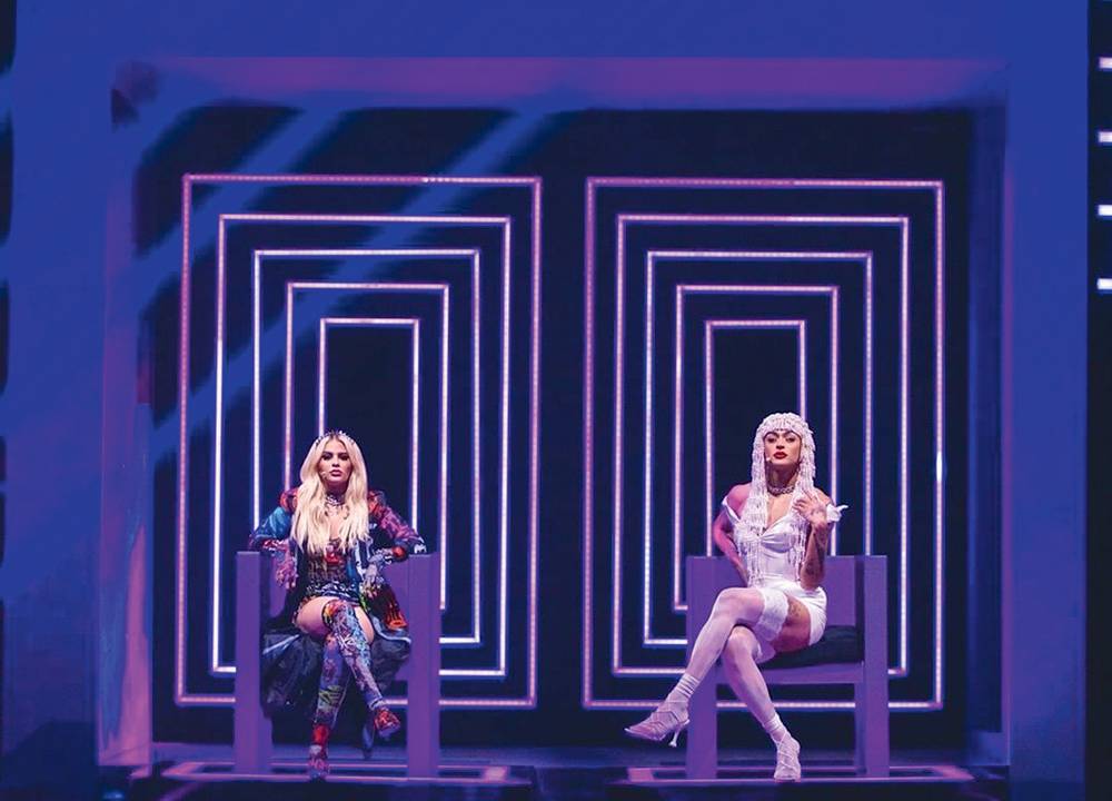 Imagem mostra palco com duas pessoas sentadas em cadeiras, em frente a fundo com projeção retangular roxa.