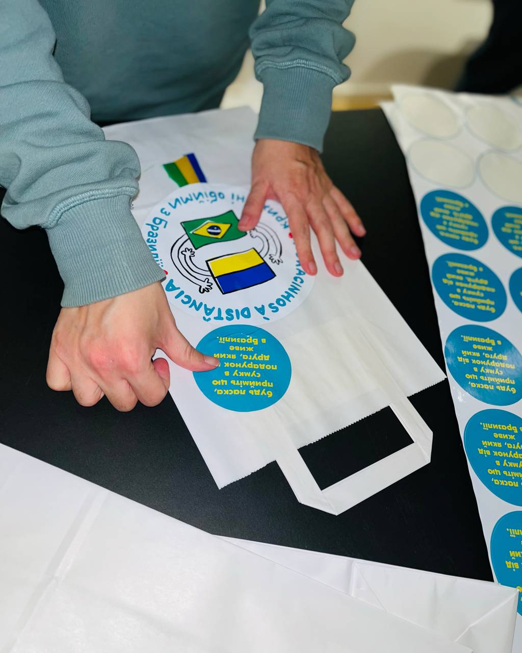 Foto exibe par de mãos preparando logotipo de sacolinha branca.