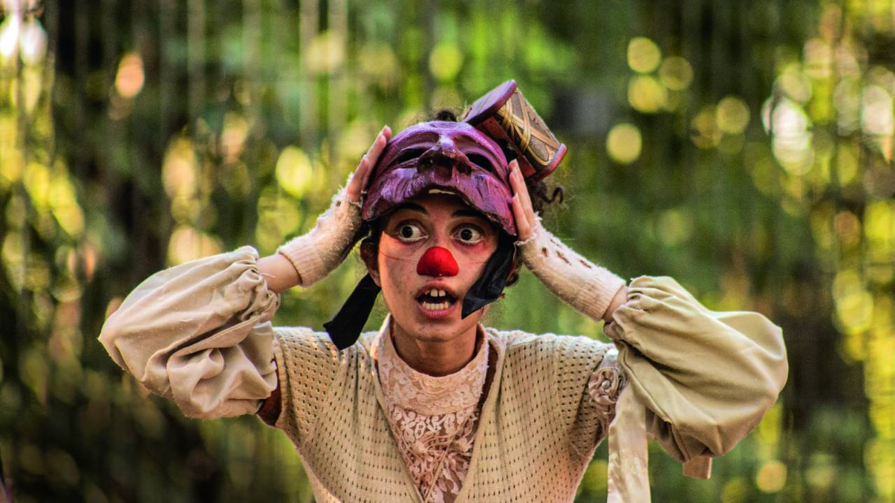 Uma atriz branca, de olhos e cabelos escuros, aparece do peito para cima com as mãos na cabeça. Ela usa um nariz de palhaço e uma máscara de teatro, puxada até a testa