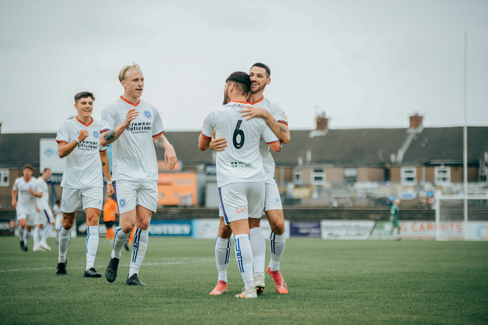 Imagem mostra jogadores de futebol sorrindo e se abraçando. Eles vestem uniformes brancos