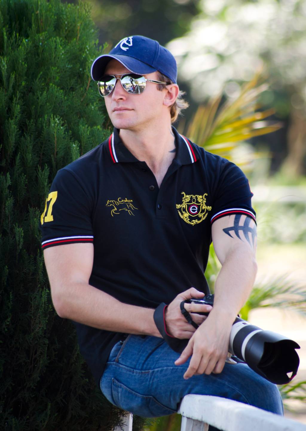 Raphael Macek (Arquivo Pessoal) Raphael Macek, fotógrafo especializado em cavalos, posa sentado com jeans, polo, óculos escuros e boné azul. Segura na mão direita sua câmera.