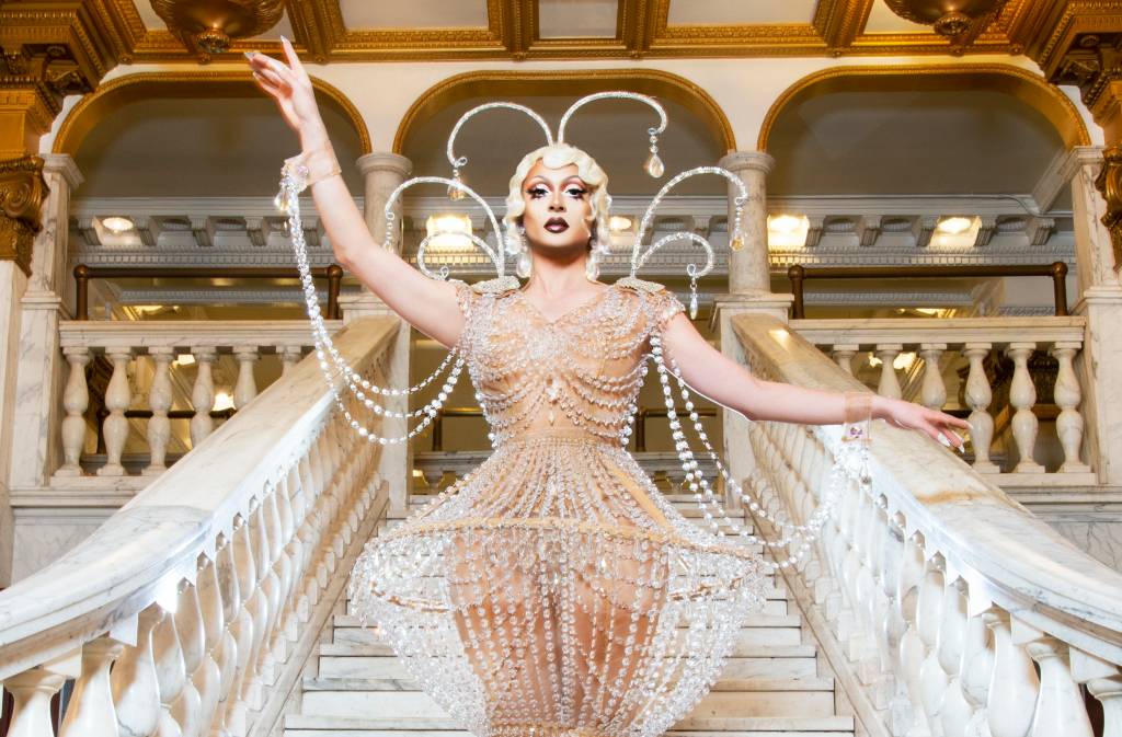 Drag queen Denali posa em escadaria branca com vestido de pedras, uma das mãos para cima, e o teto dourado em evidência.