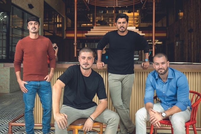 Quatro homens brancos e magros, sócios do Vila Anália, posam em frente ao estabelecimento. Dois estão sentado, à frente, e outros dois estão em pré, atrás. O ambiente é iluminado por luz amarela atrás.