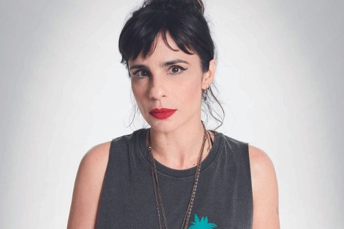 Maria Ribeiro é uma mulher em seus 40 e poucos anos. É branca, magra, de cabelos pretos e lisos amarrados em um coque. Usa batom vermelho e blusa cinza. Posa do peito para cima em um fundo branco