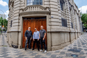 Imagem mostra três homens em frente a porta de madeira