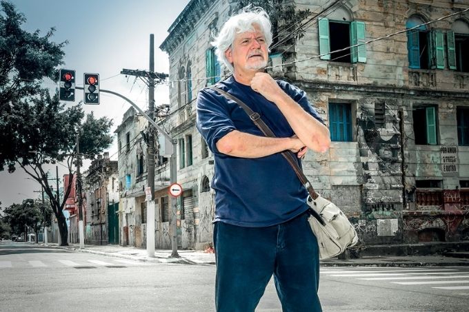 Edu é um homem branco, idoso de cabelos grisalhos. Ele usa uma camisa azul marinho, uma calça jeans escura e uma bolsa lateral cinza. Posa em pé no meio da Rua Helvétia, onde costumava ser a Cracolândia