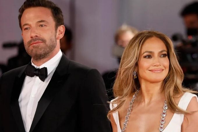 Ben Affleck, um homem magro, branco e de barba e cabelos escuros, usa um terno e posa ao lado de sua noiva, Jennifer Lopez, uma mulher de pele morena e cabelos lisos, longos e castanhos. Ela usa um vestido branco com pedras prateadas e um amplo decote em U.