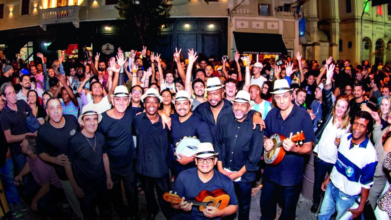 Imagem mostra aglomeração de pessoas em torno de grupo de samba com instrumentos, na rua