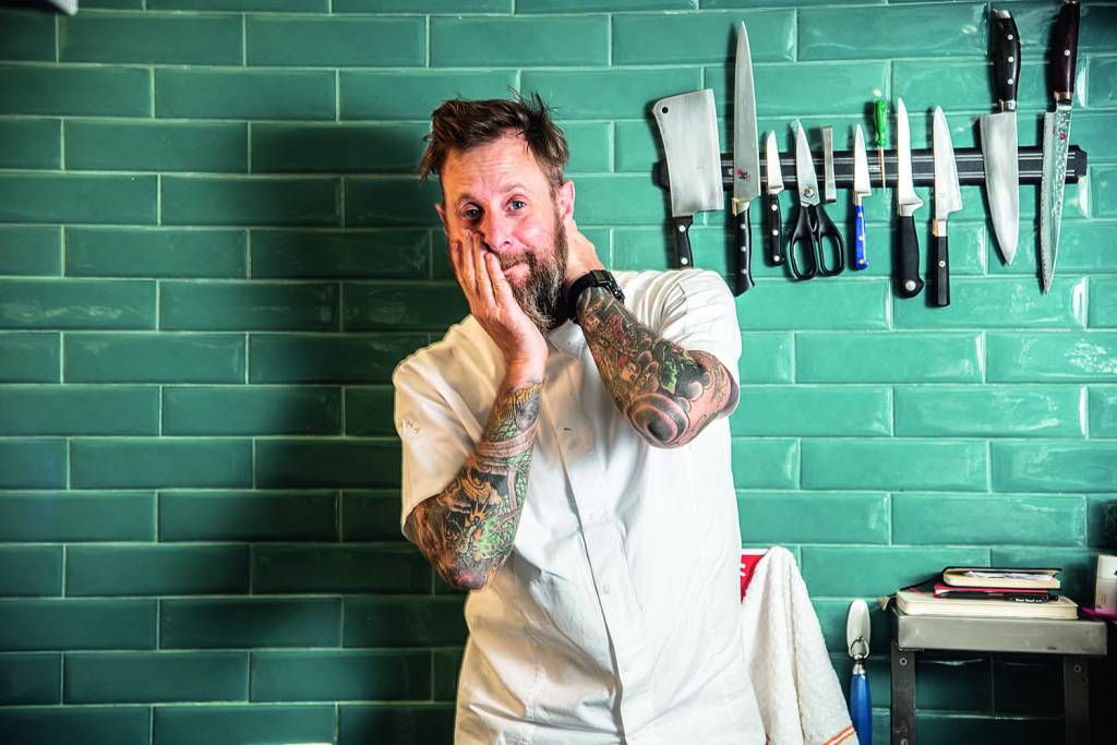 Chef André Mifano posa vestindo avental em frente à parede de azulejos de cor azul. Na mesma parede, há um faqueiro pendurado à direita de Mifano.