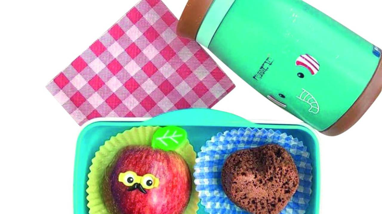 Pote verde água com uma maçã e um bolinho de chocolate em formato de coração dentro. A maçã tem um pino de plástico que simula um óculos e um bigode. Ao lado, há um guardanapo quadriculado vermelho e uma garrafa com estampa de bichinho
