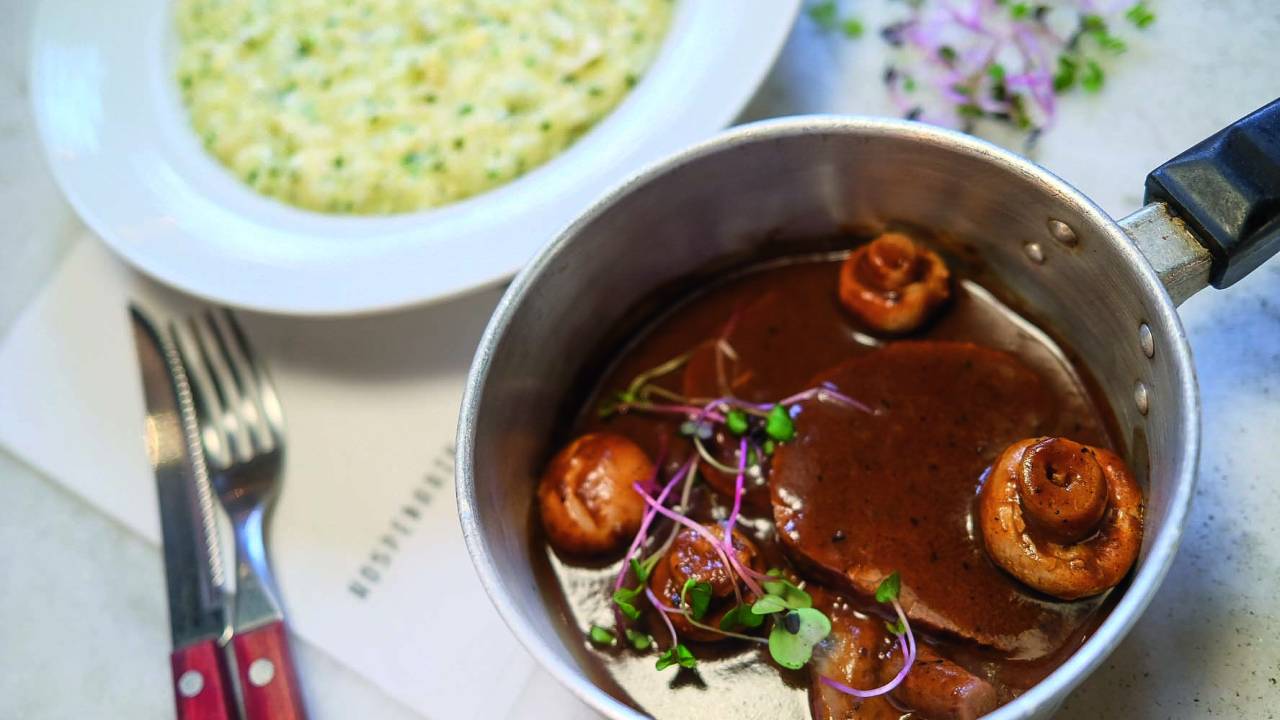 Carne com molho servida em pequena panela metálica e, ao fundo, seu acompanhamento, o arroz cremoso, servido em um prato fundo de louça branca