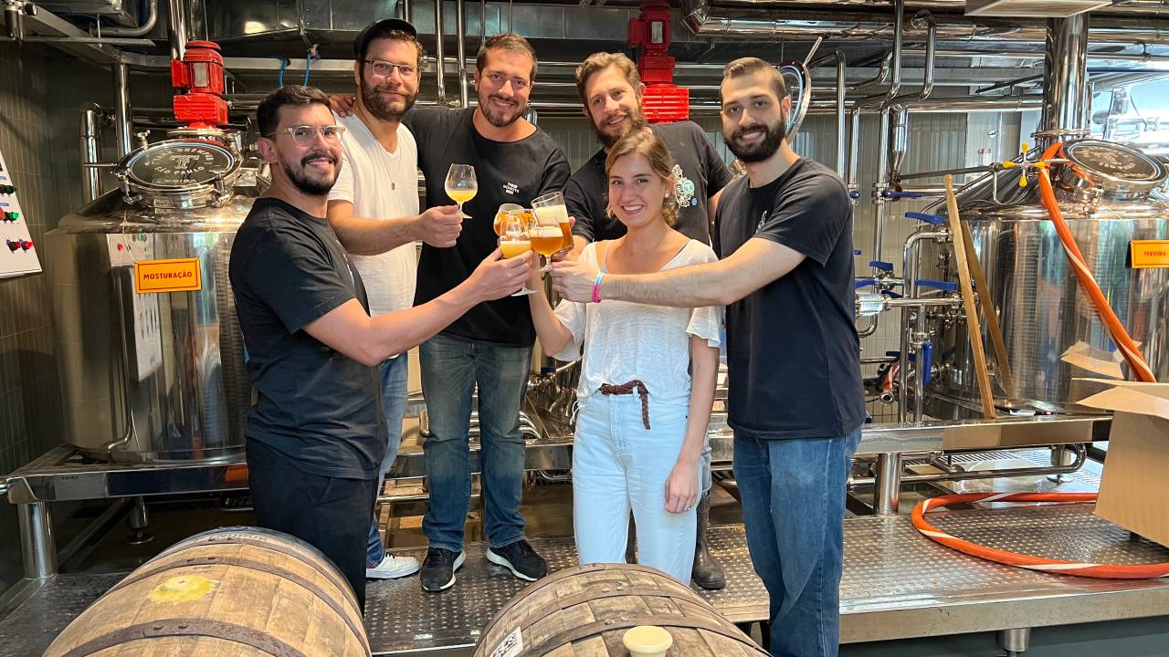 Cinco homens (quatro deles com camisetas pretas e um vestindo uma camiseta branca) posam junto de uma mulher segurando copos de cerveja em frente a tanques da bebida