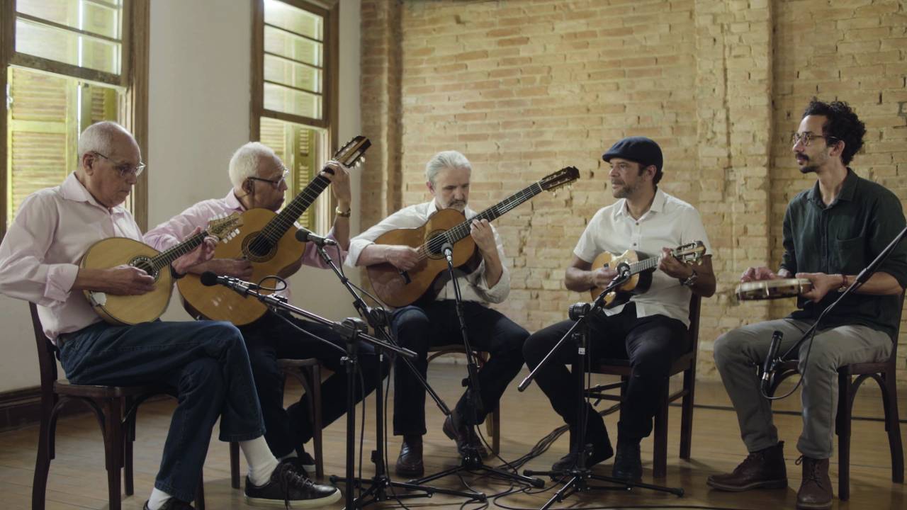 Imagem mostra cinco homens sentados em meia lua com instrumentos nas mãos. Eles vestem camisas claras e calças escuras