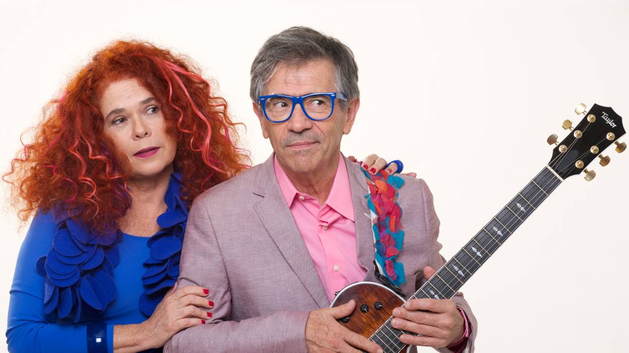 Sandra e Paulo aparecem uma ao lado do outro, com ela se apoiando em um dos braços dele. Ela é uma mulher branca, de longos cabelos ruivos e cacheados. Ele é um senhor branco de cabelos grisalhos. Usam roupas coloridas e ele segura uma guitarra