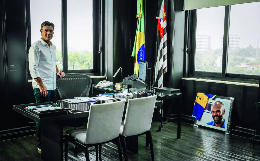RODRIGOGARCIA_14.JPG Imagem mostra homem de camisa branca em sala com mesa, cadeiras, bandeiras do Brasil e de São Pauloe um quadro de um homem no chão, encostado na parede