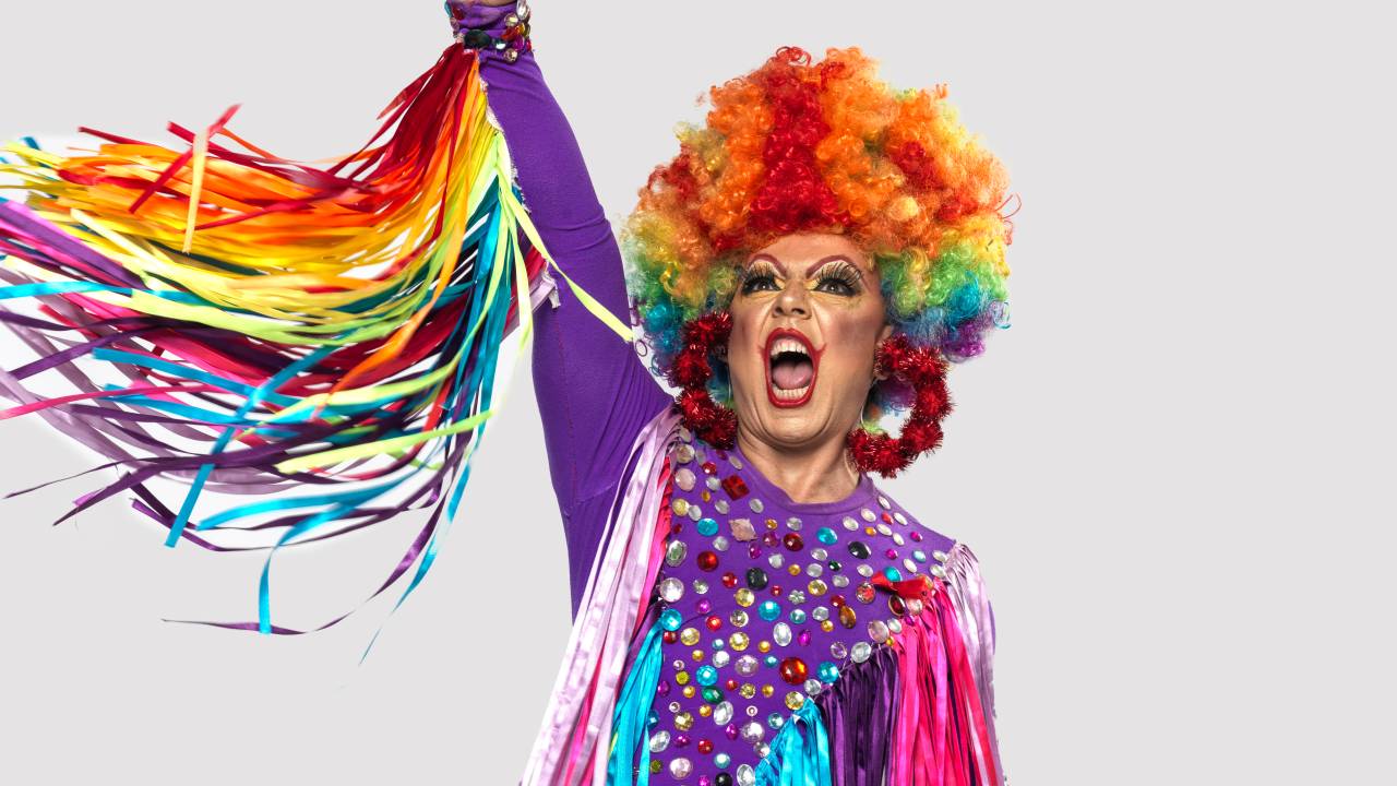 Drag queen TchaKa, personificada por Valder Bastos, posa com um braço empunhado ao alto. Veste peruca colorida com roupa roxa e adereços coloridos esvoaçantes.