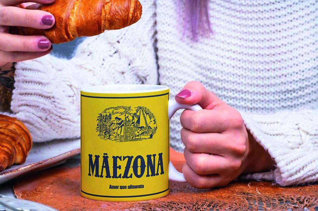 caneca Foto mostra close nas mãos de mulher branca que segura uma caneca em cima de uma mesa e um pão na outra mão. A caneca tem uma estampa amarela, que lembra a embalagem famosa de amido de milho, com os dizeres