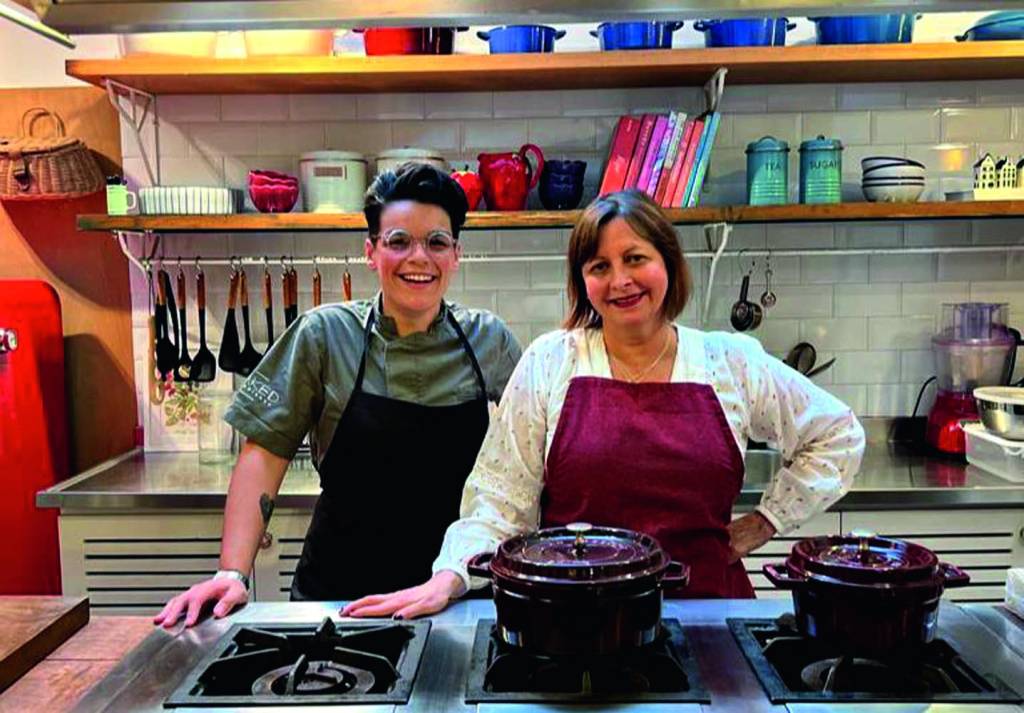 Cristiane Skrings e Carla Pernambuco posam lado a lado usando aventais de diferentes cores em um ambiente de cozinha