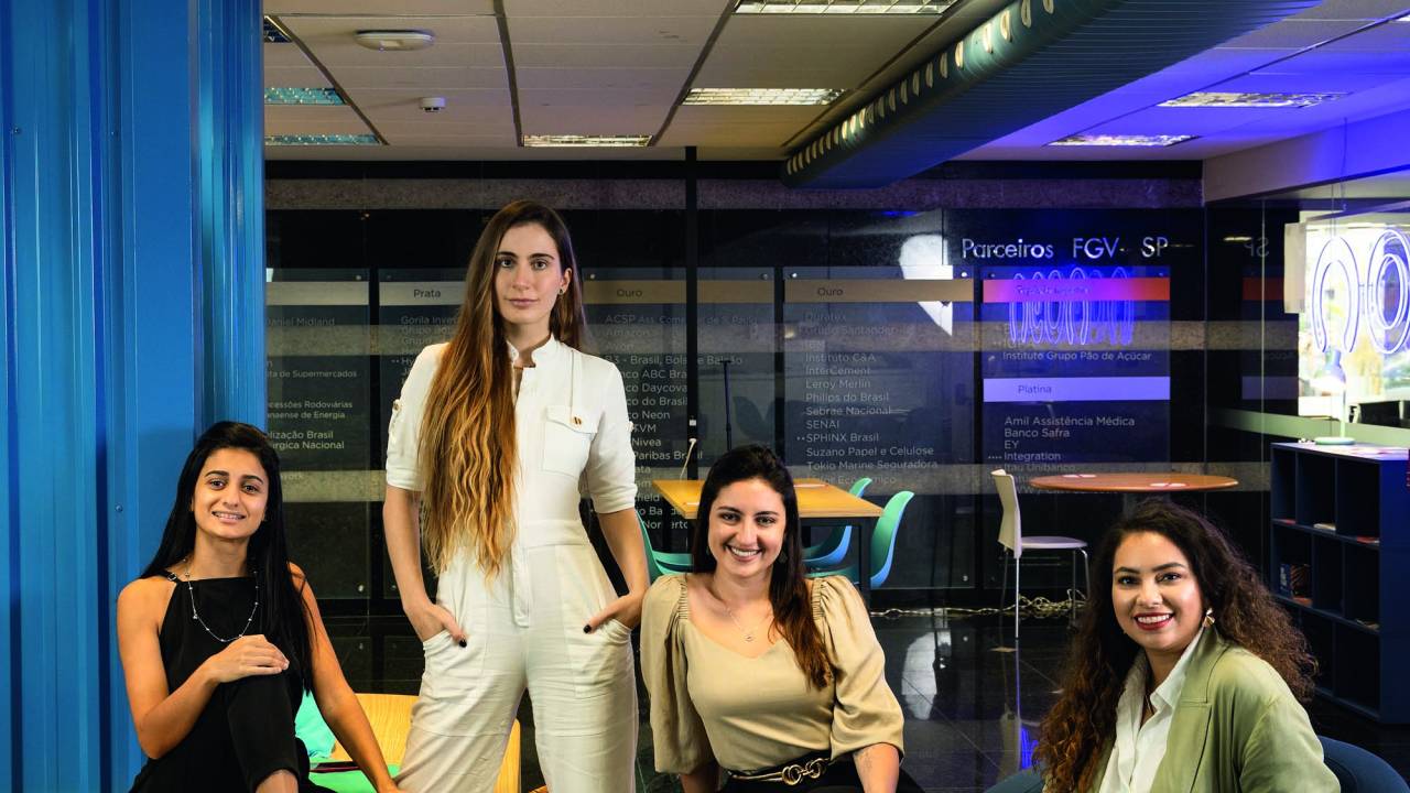 Imagem mostra quatro mulheres sorrindo. Três estão sentadas, uma está em pé. Elas estão enfileiradas.