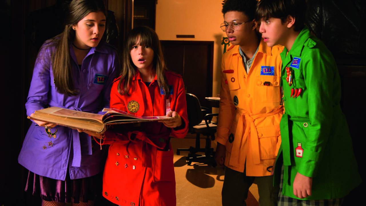 Imagem mostra quatro crianças de roupas coloridas ao redor de livro.