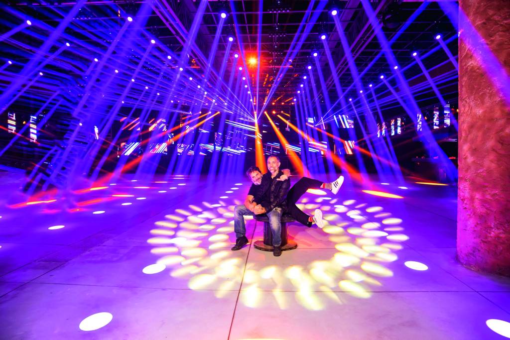 Klaus Pian e Moises Gibi — Komplexo Tempo Imagem mostra dois homens sentados em um banco de madeira no centro de um espaço grande, iluminado por luzes coloridas.