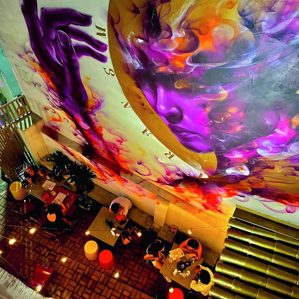 3 — (PÁGINA 2) CASA FLUIDA (DIVULGAÇÃO).jpg Imagem mostra pessoas sentadas em mesas ao lado de parede com graffiti enorme mostrando um rosto pintado de roxo.