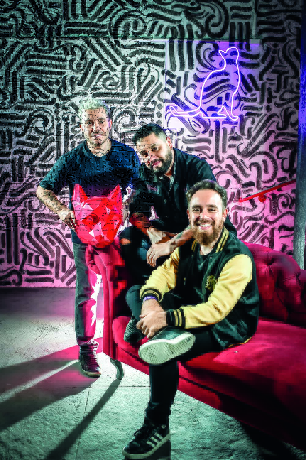 São Paulo 09 maio 2022KAT KLUB Foto Alexandre Battibugli VSP Imagem mostra trêes homens, dois em pé e um sentado, ao em espaço com parede com traços brancos e pretos ao fundo e um sofá vermelho de camurça.