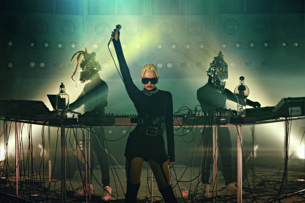 7 — Paradise Guerrilla – Camarote Pride Imagem mostramulher em palco, no centro, e dois Djs vestindo capacetes ao fundo,