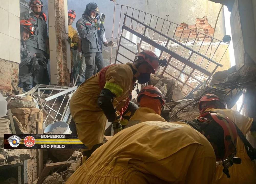 Foto exibe bombeiros em local de desabamento por explosão.